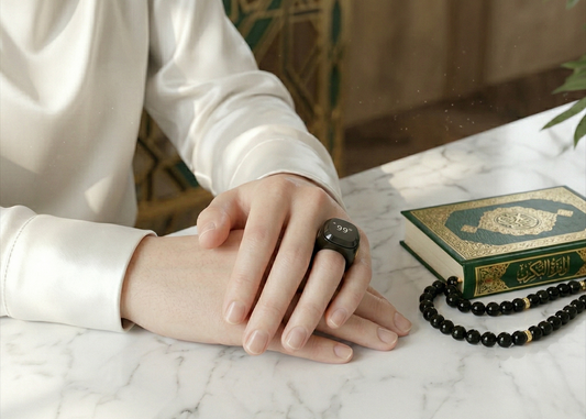 Close-up of a hand wearing a Qamar ring, resting on a marble table next to a Qur'an and prayer beads.